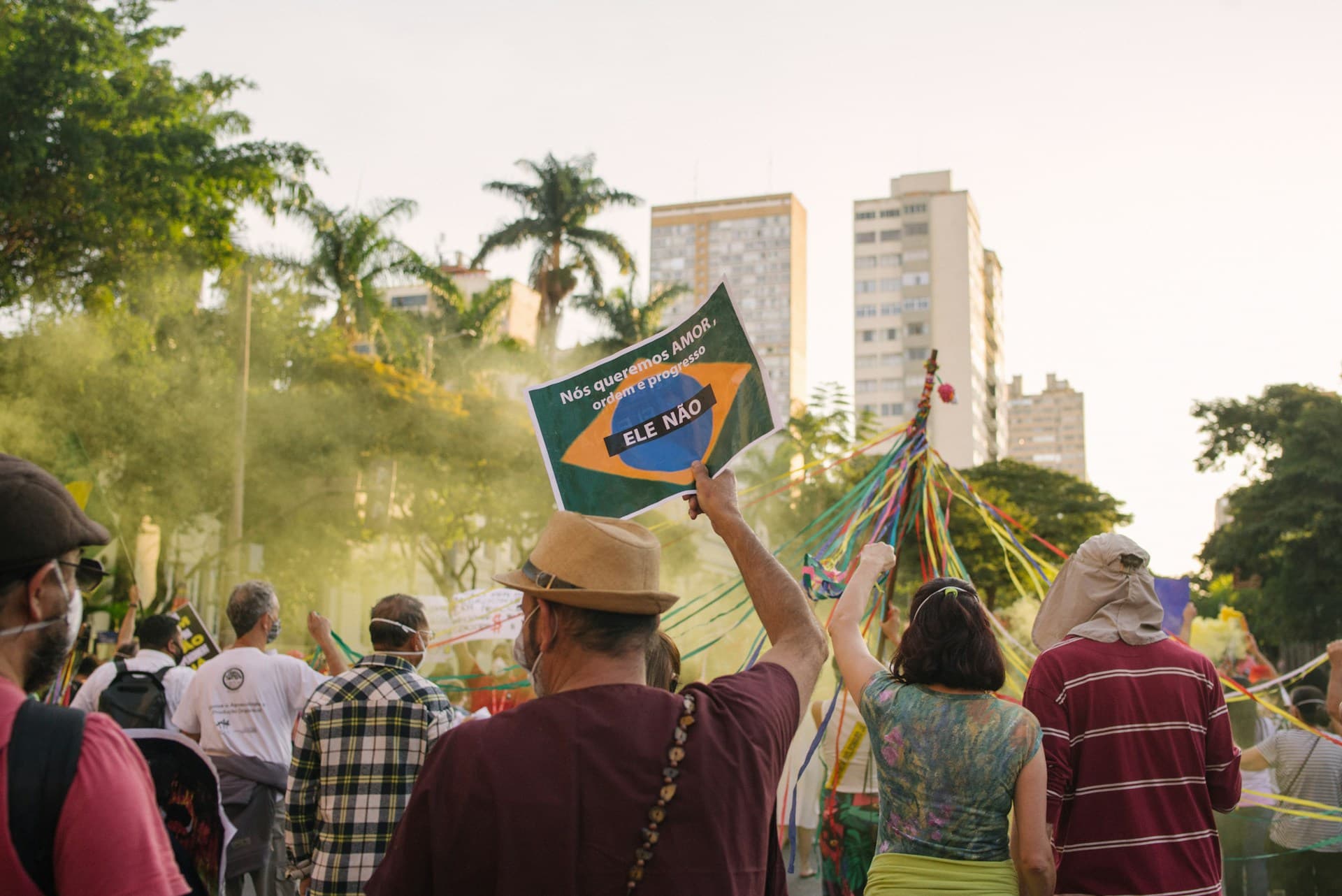 Manifestantes de máscara em uma rua com árvores e prédios, um deles erguendo um cartaz com a bandeira do Brasil